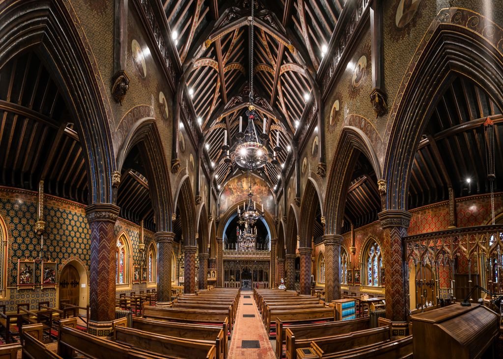 A richly coloured Gothic Revival church nave has dark timber roof trusses, pointed arcades, patterned walls and columns, stained-glass windows, wooden pews and screens leading towards the sanctuary.