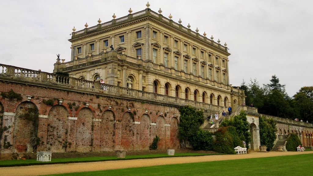 A pale Italianate country house with tall windows and roof balustrades stands high above a long brick retaining wall, arcades, stone terraces and garden stairs.