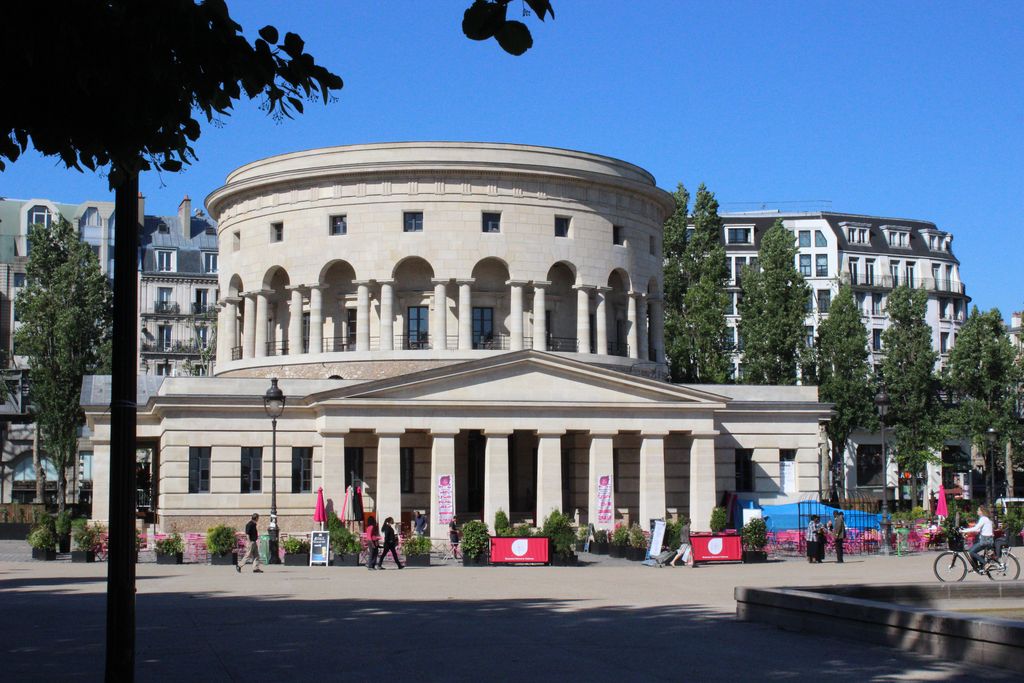 A pale stone rotunda has a square porticoed lower level and a circular upper storey surrounded by columns, with café signs, pedestrians, trees and apartment buildings around it.