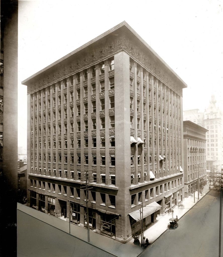 A tall early office building rises at a street corner with a heavy base, many narrow vertical window bays, richly patterned spandrels and a broad projecting cornice.