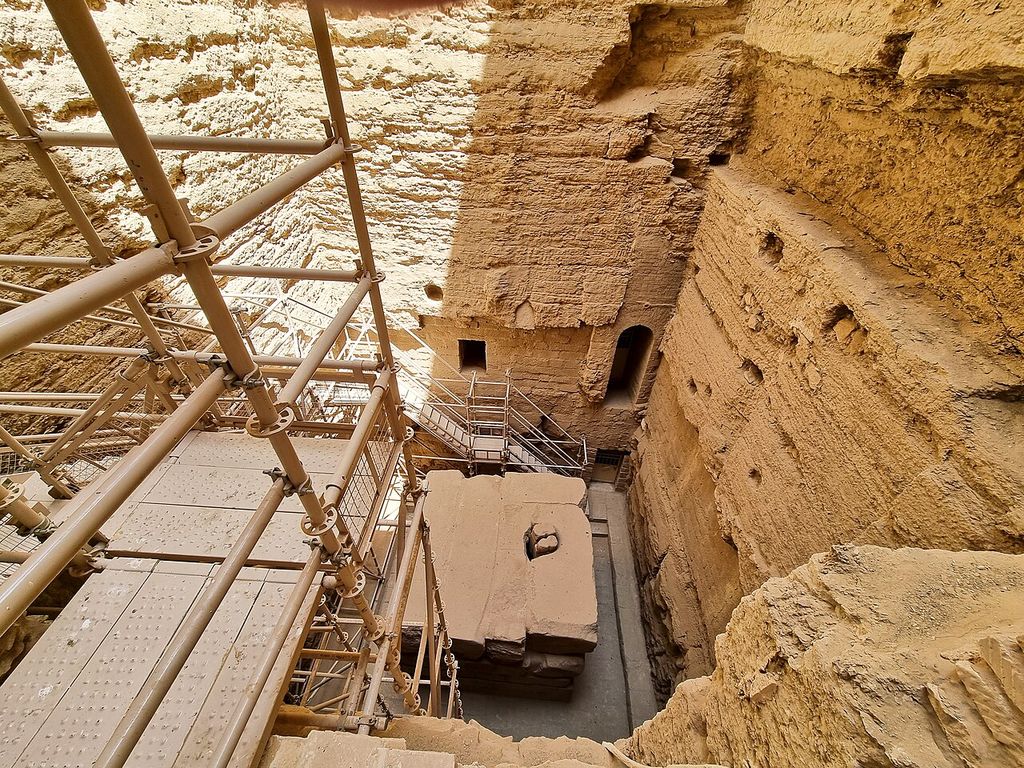 A steep view looks down past dense metal scaffolding and stairs into a deep rough-cut stone chamber with small dark openings and large blocks below.
