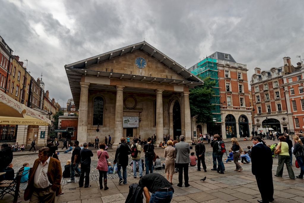 Four plain stone columns support a deep triangular pediment and clock above a church front, with crowds gathered across the busy paved square.