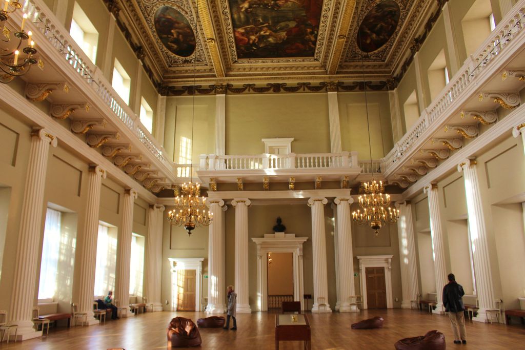 A tall ceremonial hall has giant pilasters, long upper galleries, chandeliers and large painted ceiling panels, with visitors and furniture on the floor below.