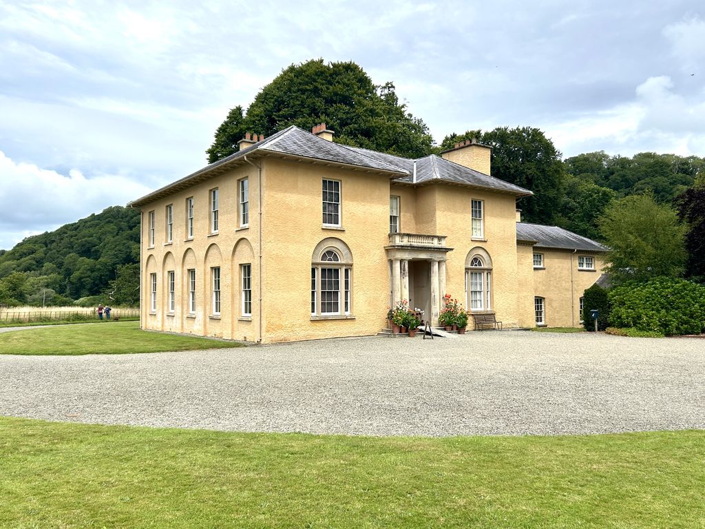 A compact cream-rendered country villa with low hipped slate roofs, tall sash windows, a small columned porch and attached side range stands beyond a broad gravel forecourt and lawn.
