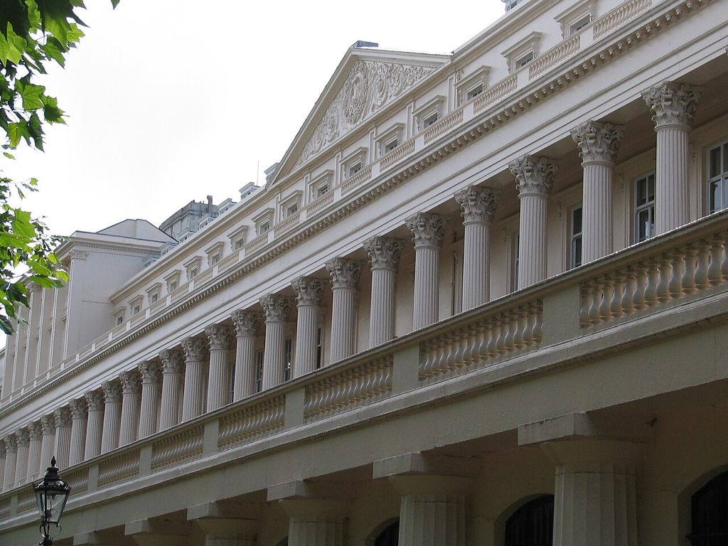 A long pale terrace recedes obliquely behind a continuous upper colonnade of fluted Corinthian columns, balustrades, repeated windows and a sculpted pediment.