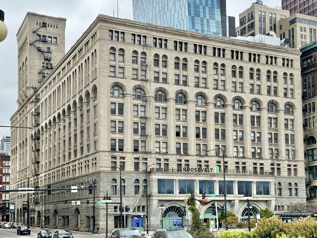 A massive pale stone Chicago block has repeated arched windows, a rusticated base and a tall office tower at one end, with modern traffic and skyscrapers around it.