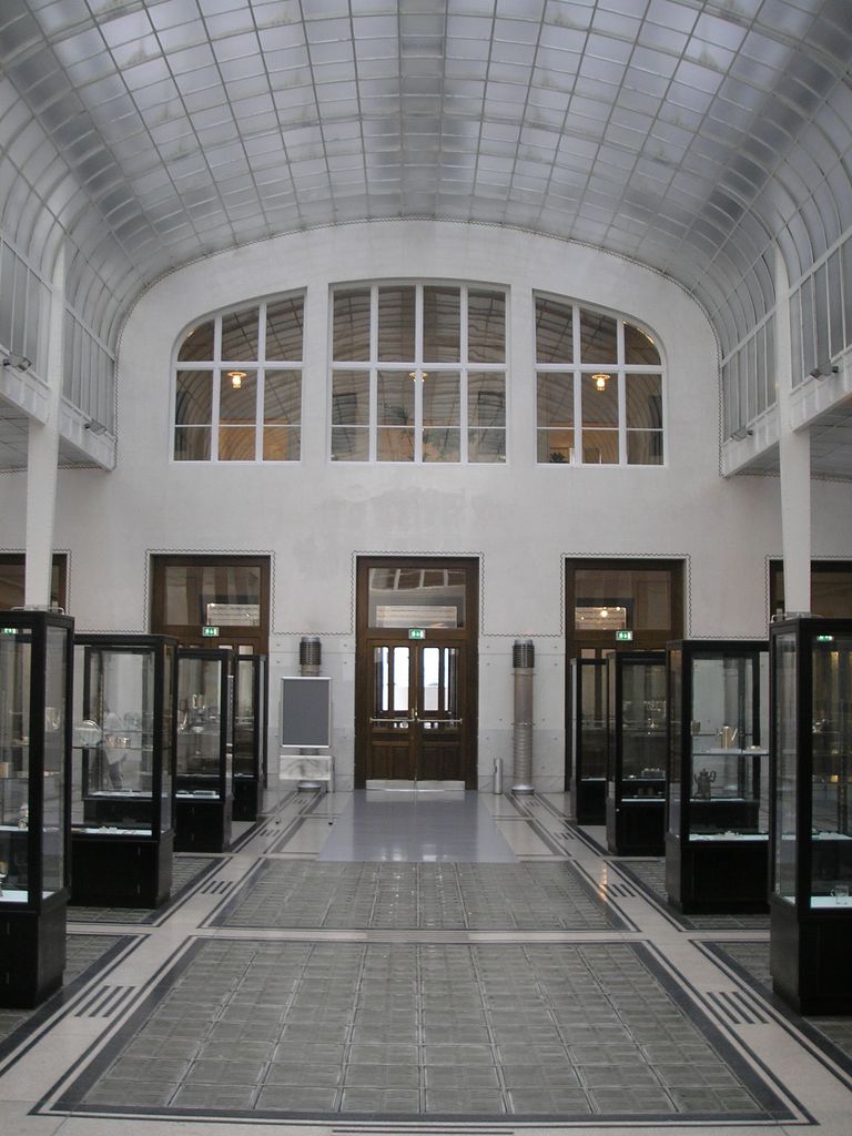 A tall white banking hall has a curved translucent glass ceiling, broad glass-block floor panels, metal-edged doorways and dark museum display cases along both sides.