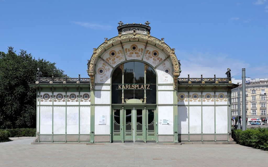 A symmetrical white pavilion has a broad arched roof, green metal framing, gilded sunflower patterns and a central entrance marked Karlsplatz under a clear blue sky.