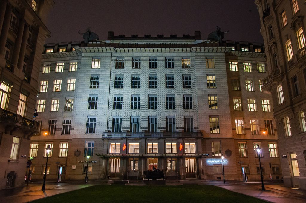 A broad pale bank façade glows at night, with regular windows and thousands of small dark points across its marble surface between older neighbouring buildings.