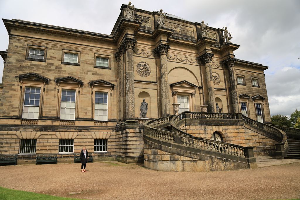 A grand stone country-house front has a projecting centre with four Corinthian columns, statues, carved panels and a wide divided stair above a rusticated basement.