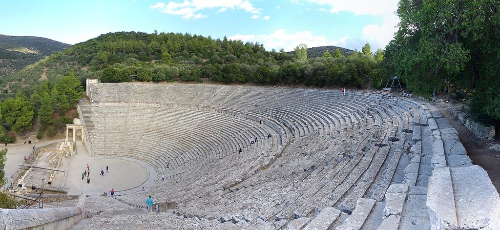 Broad concentric tiers of pale stone seats and radial aisles descend to a circular orchestra on a wooded hillside.