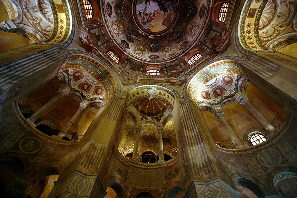 An upward wide-angle view passes tall piers and curved gallery arcades toward a richly decorated central dome, with lower spaces in shadow.