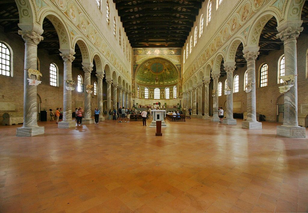 Two long arcades of marble columns divide a bright nave from side aisles beneath clerestory windows and a timber roof, ending in a mosaic apse.