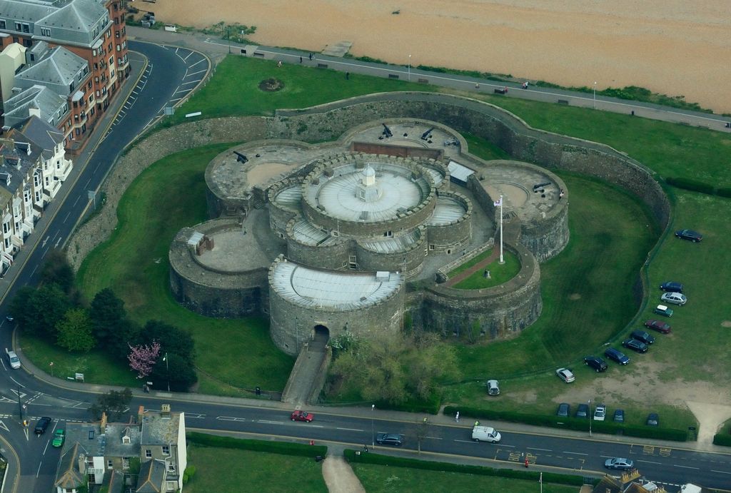 A low circular coastal fort with rounded platforms stands inside a dry ditch beside a beach and town.