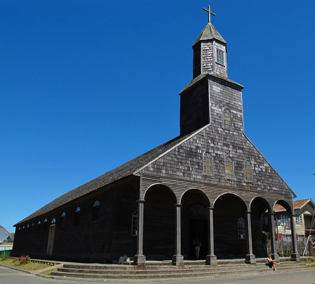 A broad dark timber-shingled church has a six-arch porch and central two-stage tower, with a seated person and timber houses beneath a vivid blue sky.