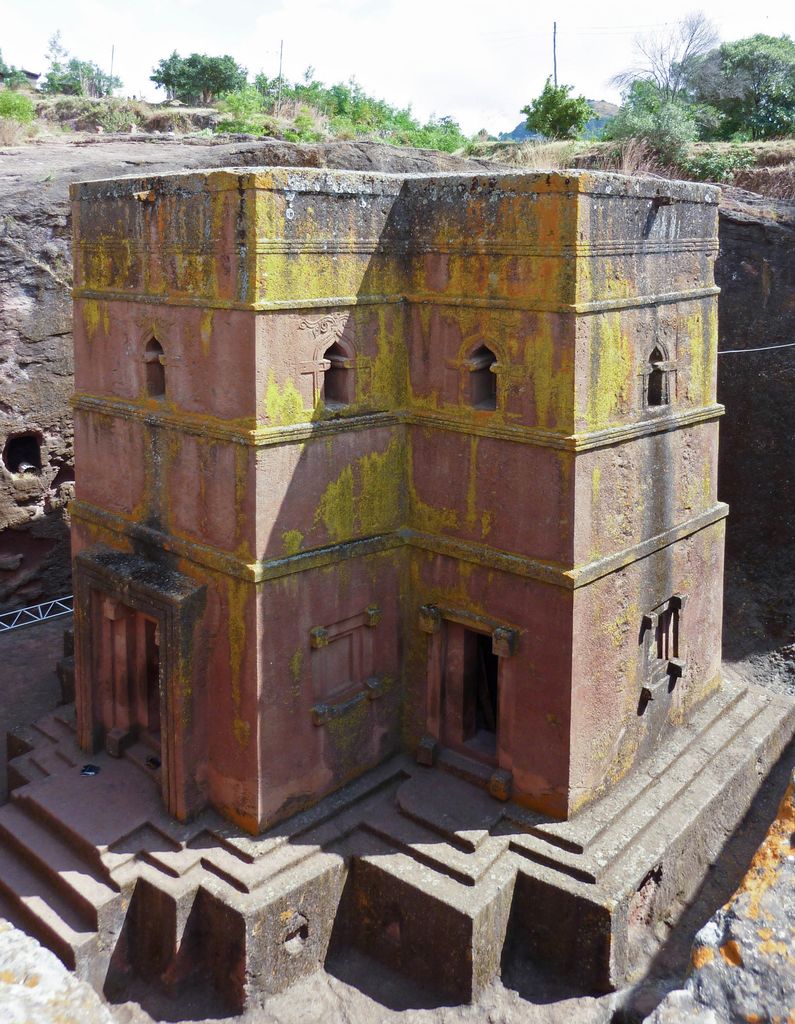 A cross-shaped reddish stone church rises from a deep rock-cut trench, with carved doors and windows, surrounding steps and living rock visible from above.