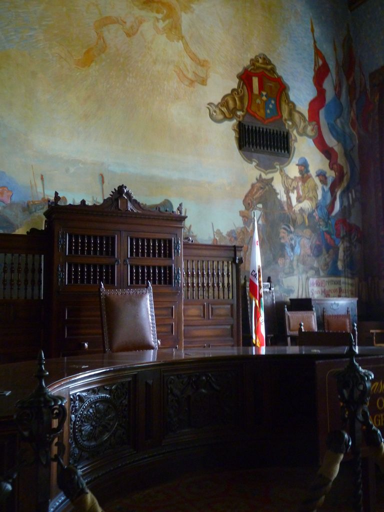 A dark carved bench and chairs stand before a large painted historical scene, crest and flags in a courthouse room.