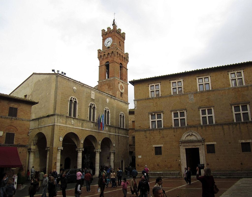 An arcaded stone town hall with a tall clock tower stands beside an ordered palazzo façade across a busy paved square.