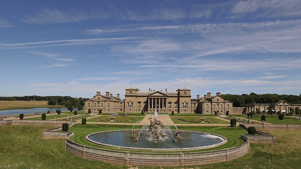 A large porticoed house and lower connected ranges stand beyond geometric lawns, clipped trees and a circular fountain.