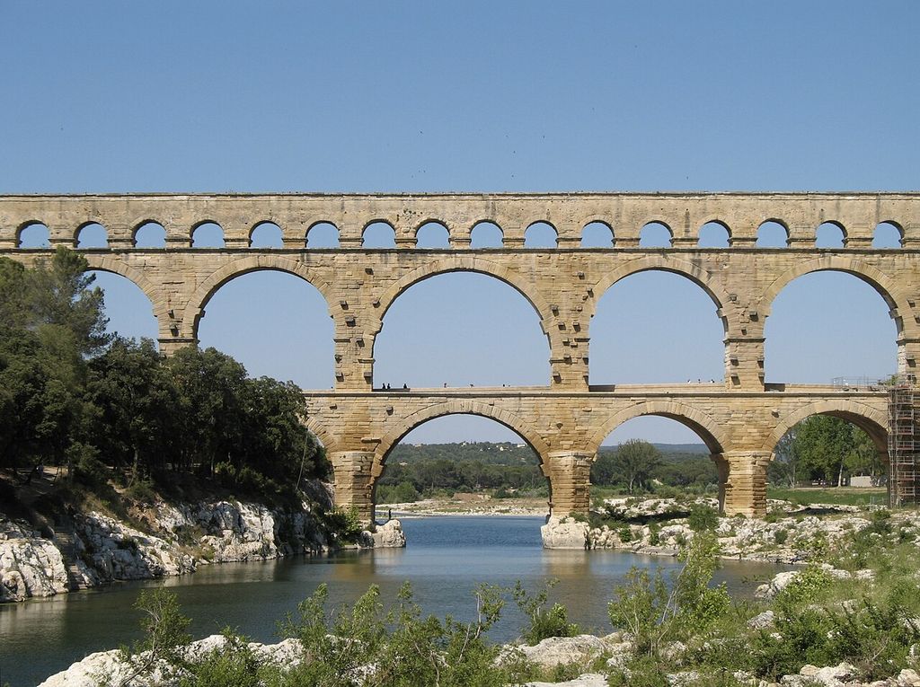 Three stacked rows of pale stone arches cross a rocky river valley, with large lower spans and many small openings along the top.