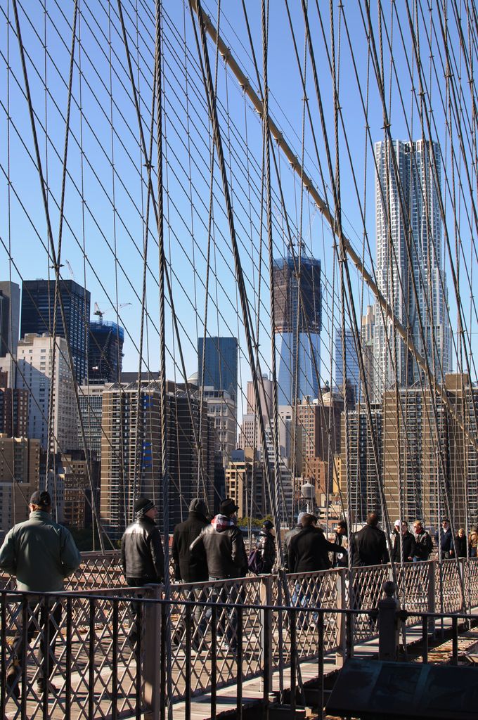 Dense vertical and diagonal cables cross a blue sky above a timber pedestrian walkway crowded with visitors and backed by city towers.