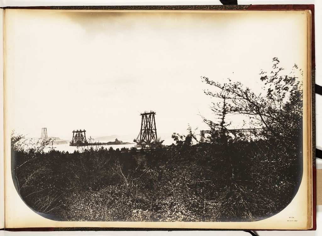 A sepia album photograph shows three distant incomplete bridge towers and approach work across water behind dark foreground foliage.