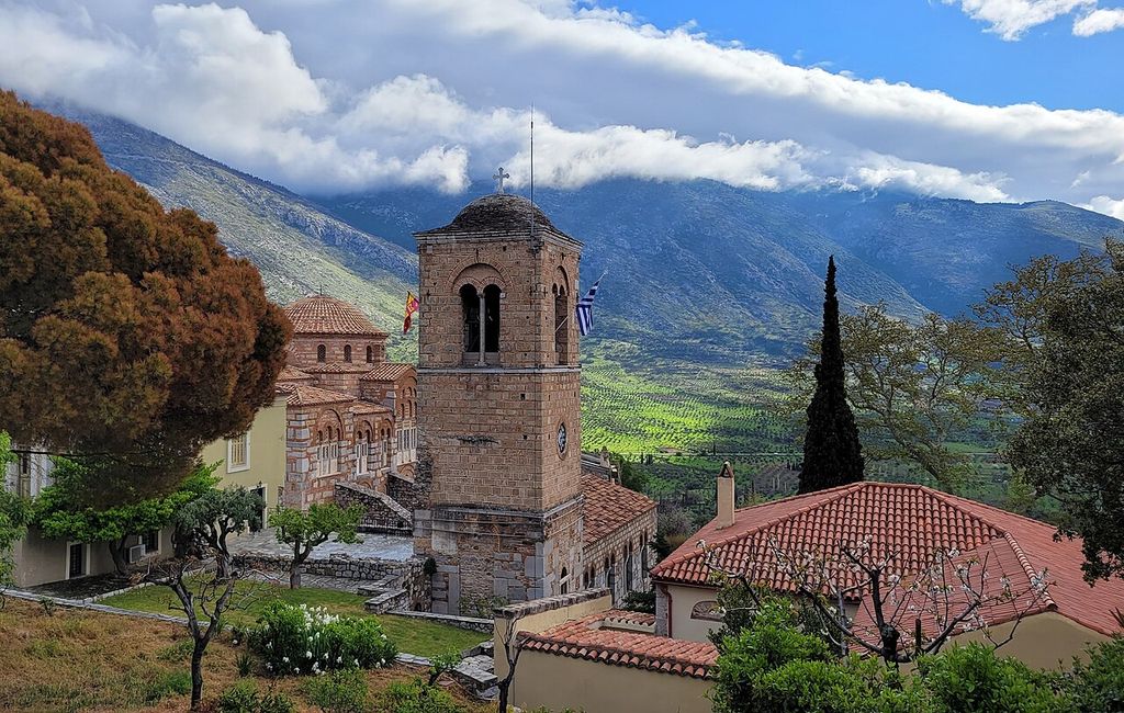 A domed church, square bell tower and tiled monastic ranges stand among trees before steep cultivated mountains under low cloud.