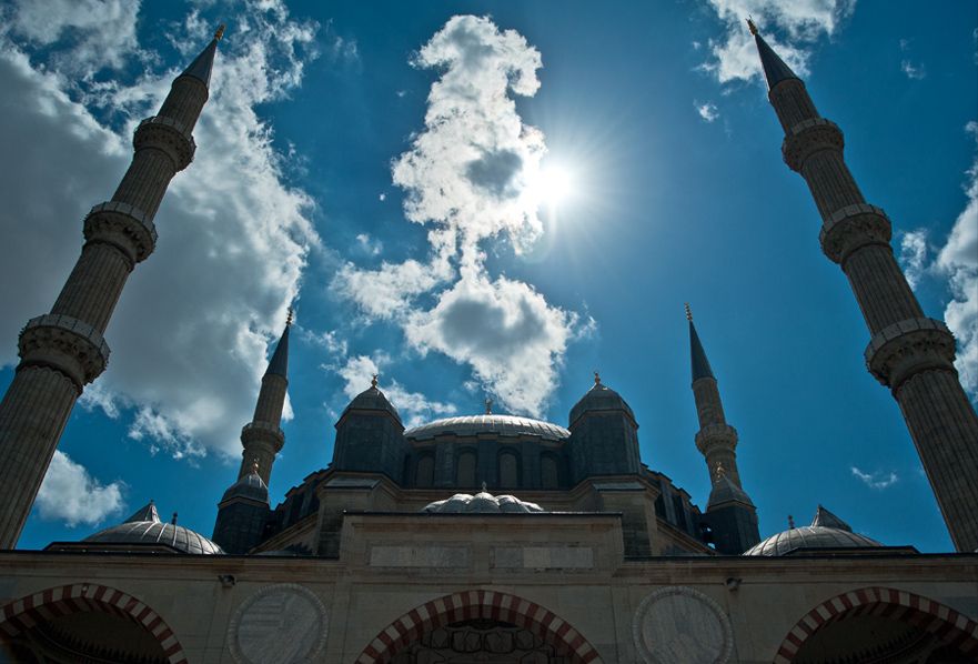 Four tall minarets frame a central leaded dome and subsidiary volumes above a courtyard arcade under bright clouds.