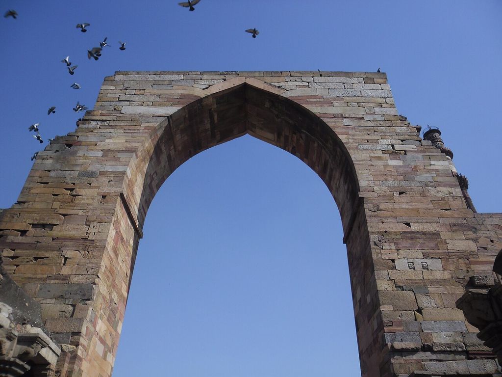 A tall pointed opening rises through horizontally coursed multicoloured stone against blue sky, with pigeons crossing above.
