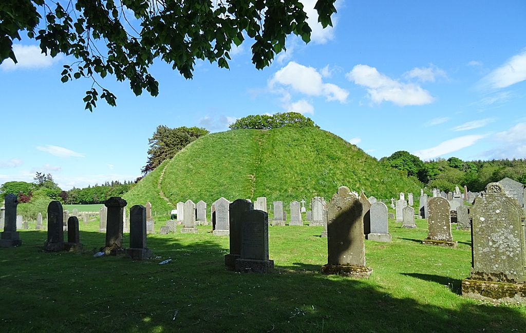 A steep grass-covered mound with trees above stands behind cemetery headstones.