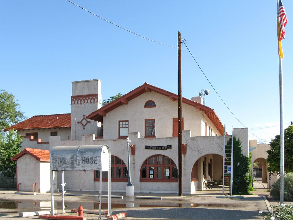 A two-storey stucco building with tiled roofs, arched windows and a shaded arcade stands beside a road under a clear blue sky.