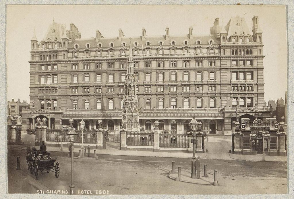 A sepia street photograph shows a large multi-storey railway hotel behind a Gothic memorial, railings, carriage and small station signs.