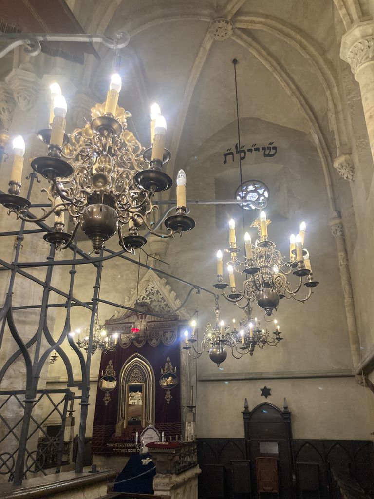 A stone synagogue interior contains an iron bimah enclosure, distant ark, ribbed vaults, carved corbels and many glowing chandeliers.