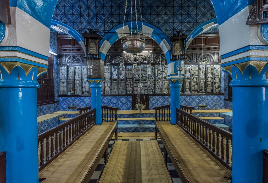 Blue columns and striped arches frame silver-fronted Torah-ark cabinets beyond rows of wooden benches and narrow passages.