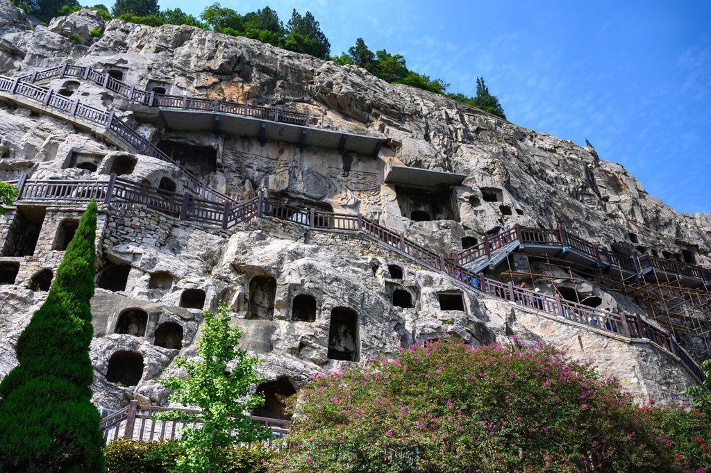 Hundreds of caves and sculpted niches pierce a steep limestone cliff crossed by modern stairs, rails, platforms and protective scaffolding.