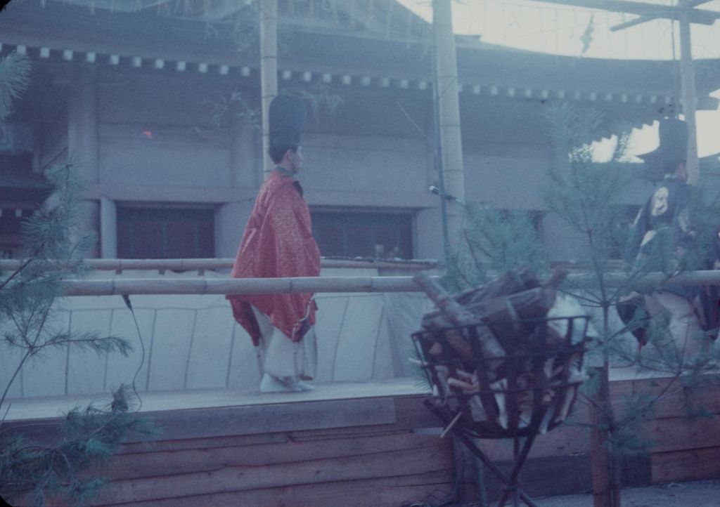 Two costumed performers stand on a raised roofed timber stage behind a low rail, with posts, open background and pine branches visible through faded colour.