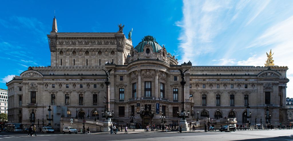 An ornate stone opera-house wing with a domed projecting pavilion, arched ground floor, high roof masses and curving ramp borders a busy street.