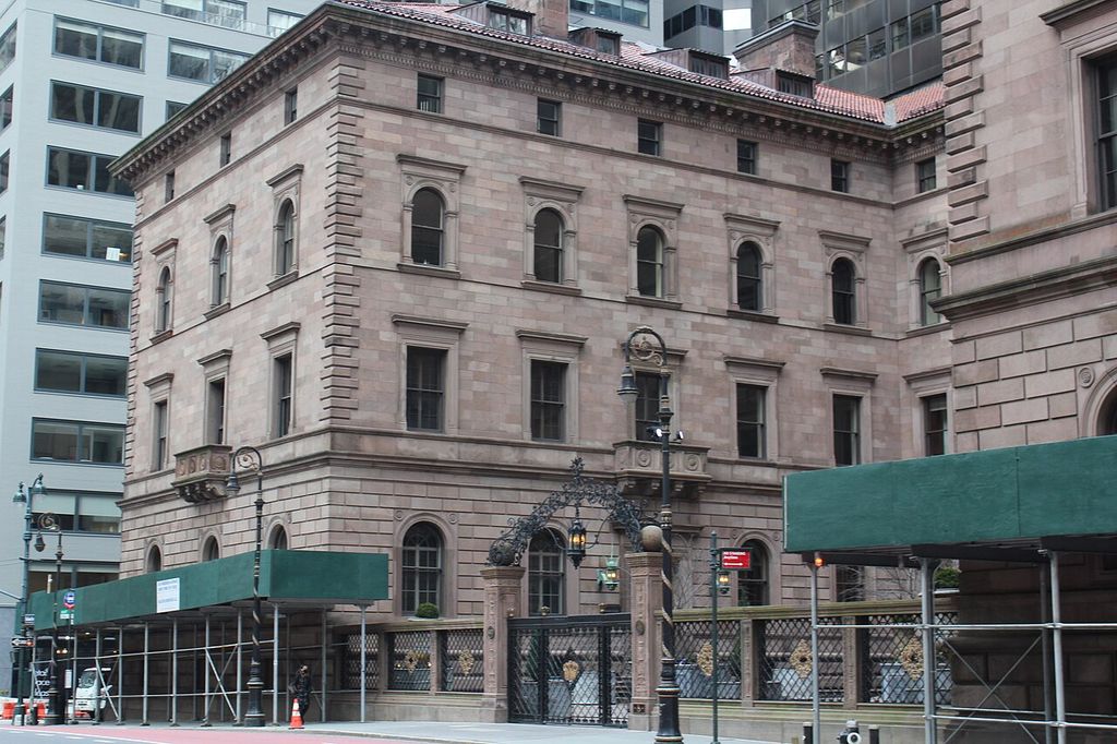 A rusticated stone palazzo-like wing with arched and rectangular windows stands behind sidewalk scaffolding and ornate gates among taller modern buildings.