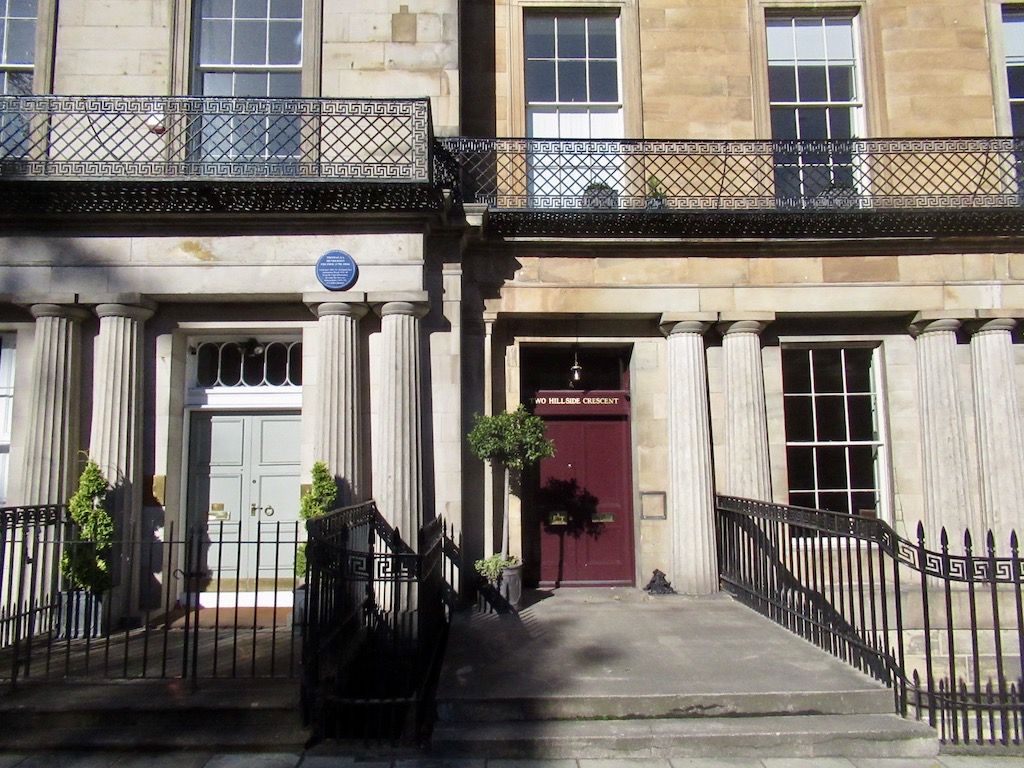 Paired fluted columns frame two townhouse doors beneath continuous balconies and railings decorated with a Greek-key pattern.