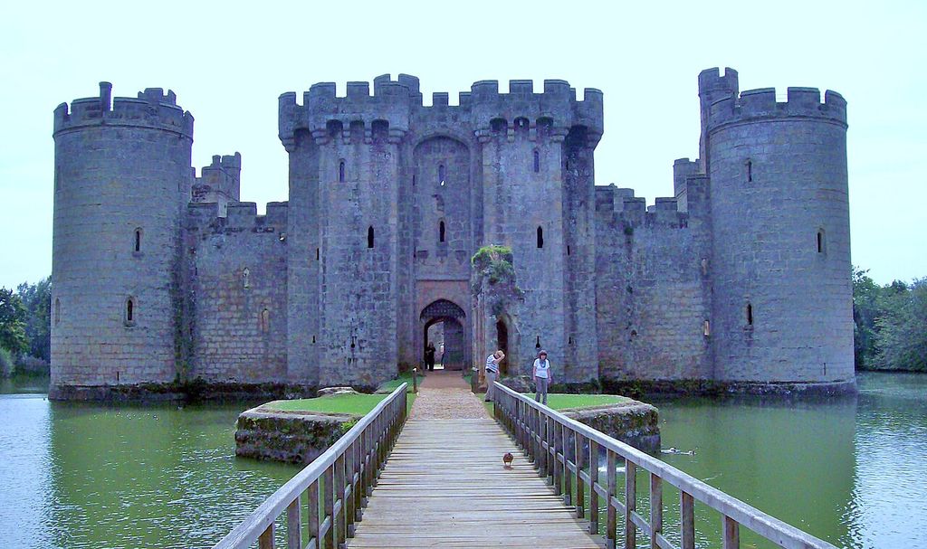 A timber footbridge crosses a broad moat toward a stone gatehouse flanked by round crenellated towers.