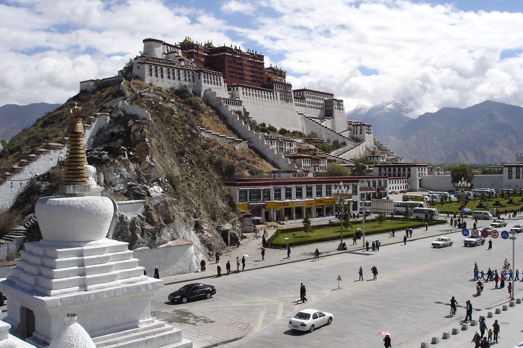 A huge white and red palace complex steps up a rocky mountain above roads, smaller buildings and distant peaks.