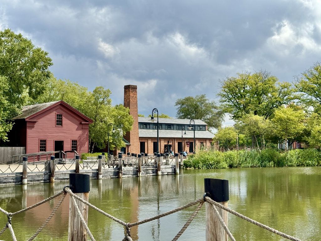 Across a pond, a red-brick workshop has a pitched roof topped by a long narrow raised monitor with dark side openings and a shallow roof of its own.