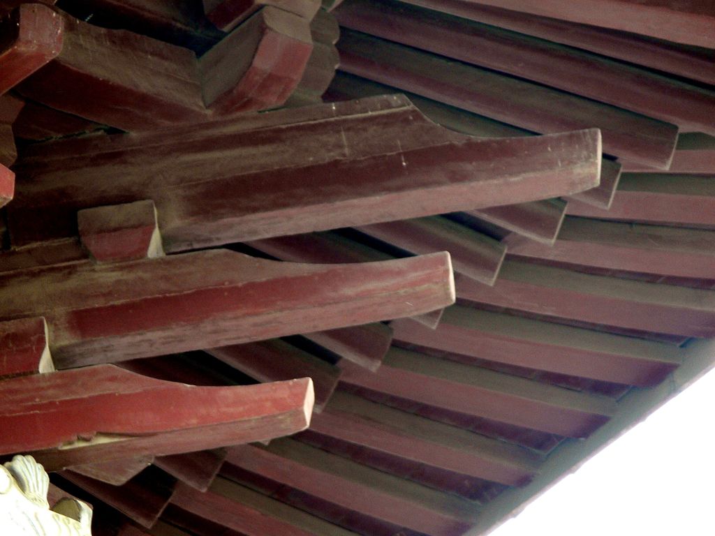 Several layers of red-painted wooden arms and blocks step outward beneath roof boards in a close upward view.