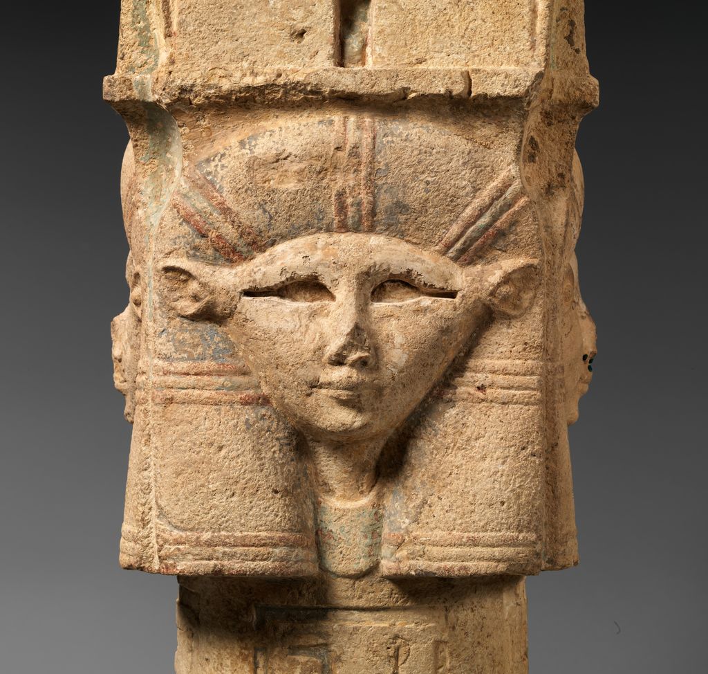 A weathered limestone capital shows Hathor's face below a block-like crown, with faded blue-green and red pigment around the hair and headdress.