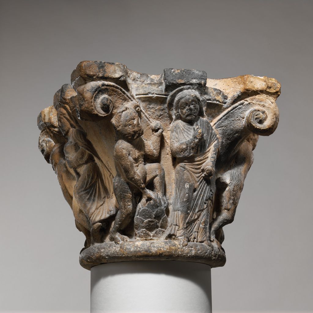 Christ and other figures are carved across the flaring face of a weathered stone capital between large spiral corner forms.