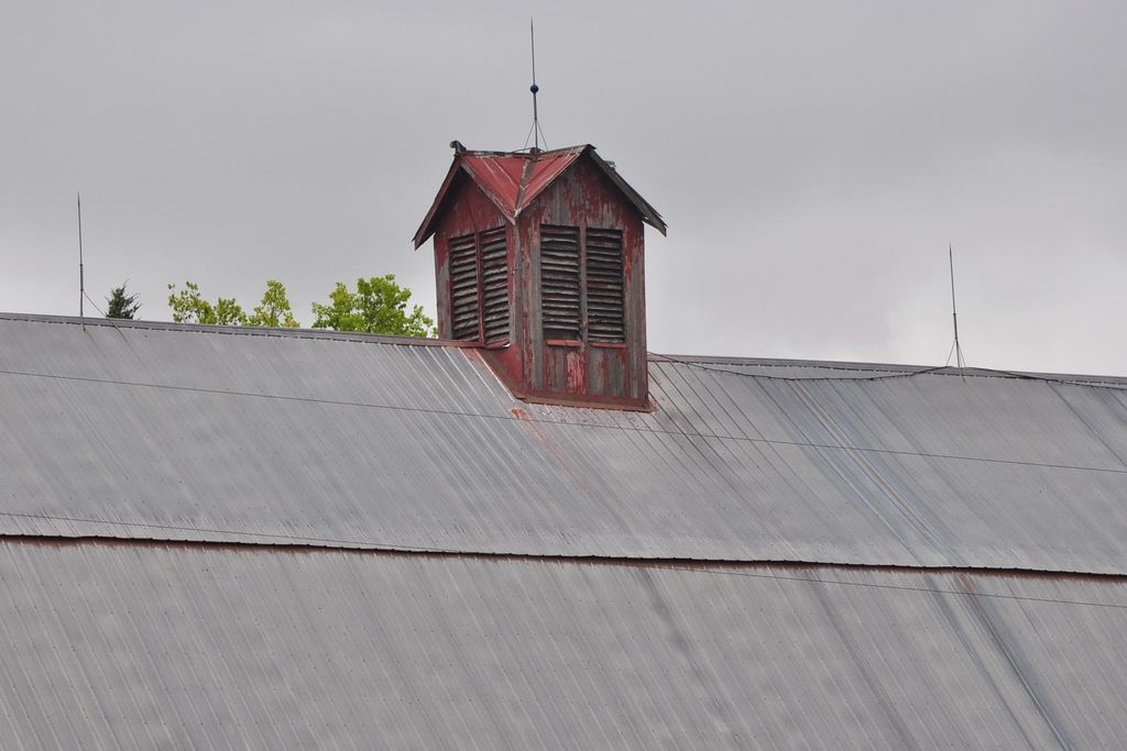 A weathered red wooden cupola with paired arched louvres and a small intersecting metal roof stands above a broad grey barn roof.