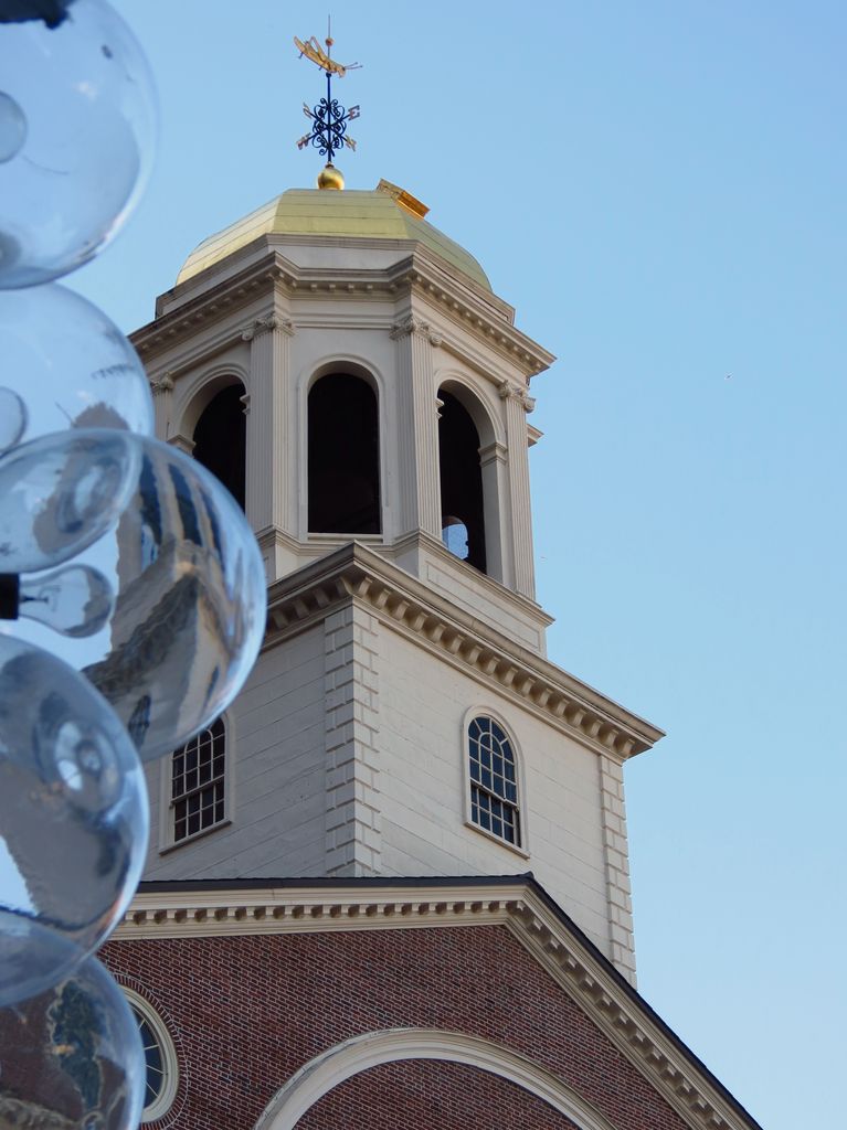A white clock tower rises to an open arched cupola, shallow metal roof and ornate grasshopper weather vane above red brick.