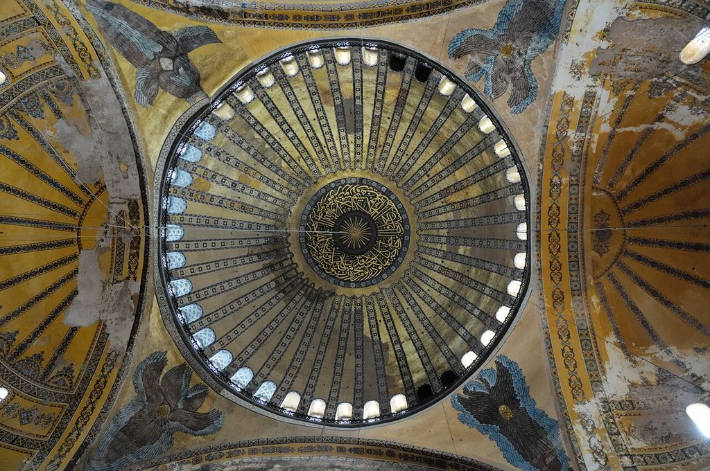 A circular gold-toned dome with radiating bands and Arabic calligraphy is encircled by bright windows above decorated curved corner surfaces.