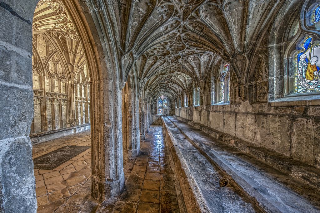A long stone washing trough runs beneath a richly vaulted cloister passage with stained-glass windows and worn paving.