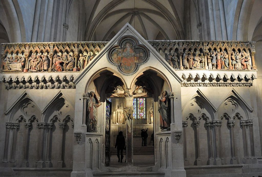 A pale stone choir screen combines two pointed doorways, painted sculpture, blind foiled arcades and small open tracery beneath a vaulted church interior.